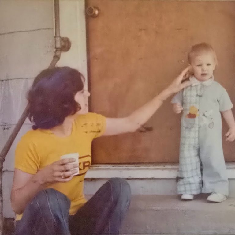 Vintage photo of a toddler in plaid overalls standing on a porch step while their mother gently touches their ear. The moment captures early signs of undiagnosed Ehlers-Danlos Syndrome and the quiet tenderness of caregiving in a small-town 1970s setting.