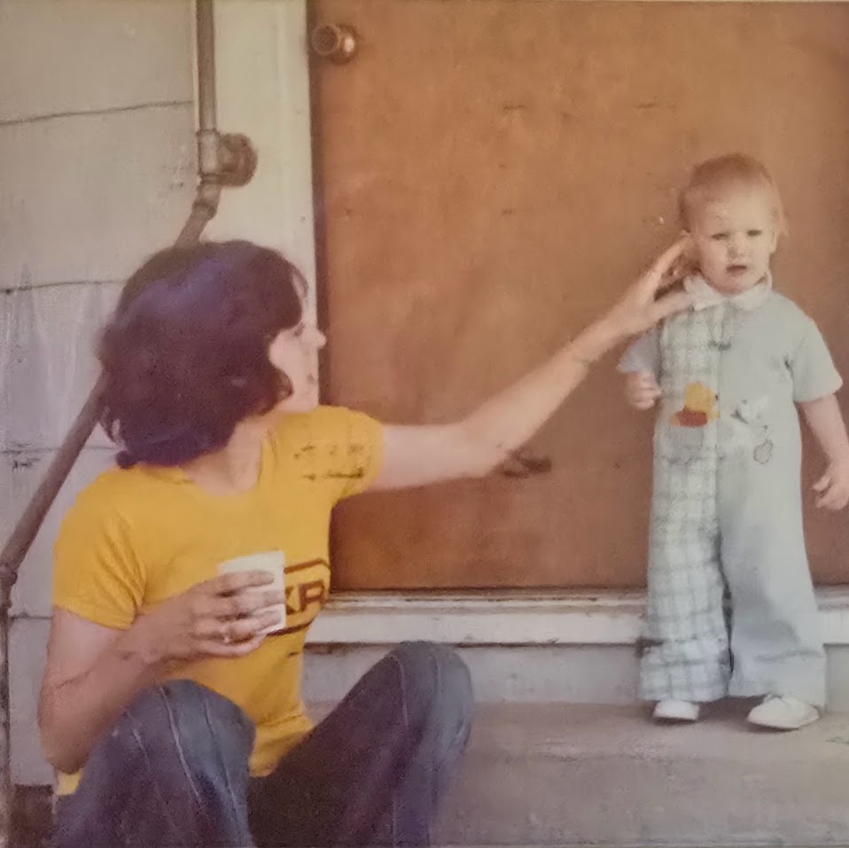 Vintage photo of a toddler in plaid overalls standing on a porch step while their mother gently touches their ear. The moment captures early signs of undiagnosed Ehlers-Danlos Syndrome and the quiet tenderness of caregiving in a small-town 1970s setting.