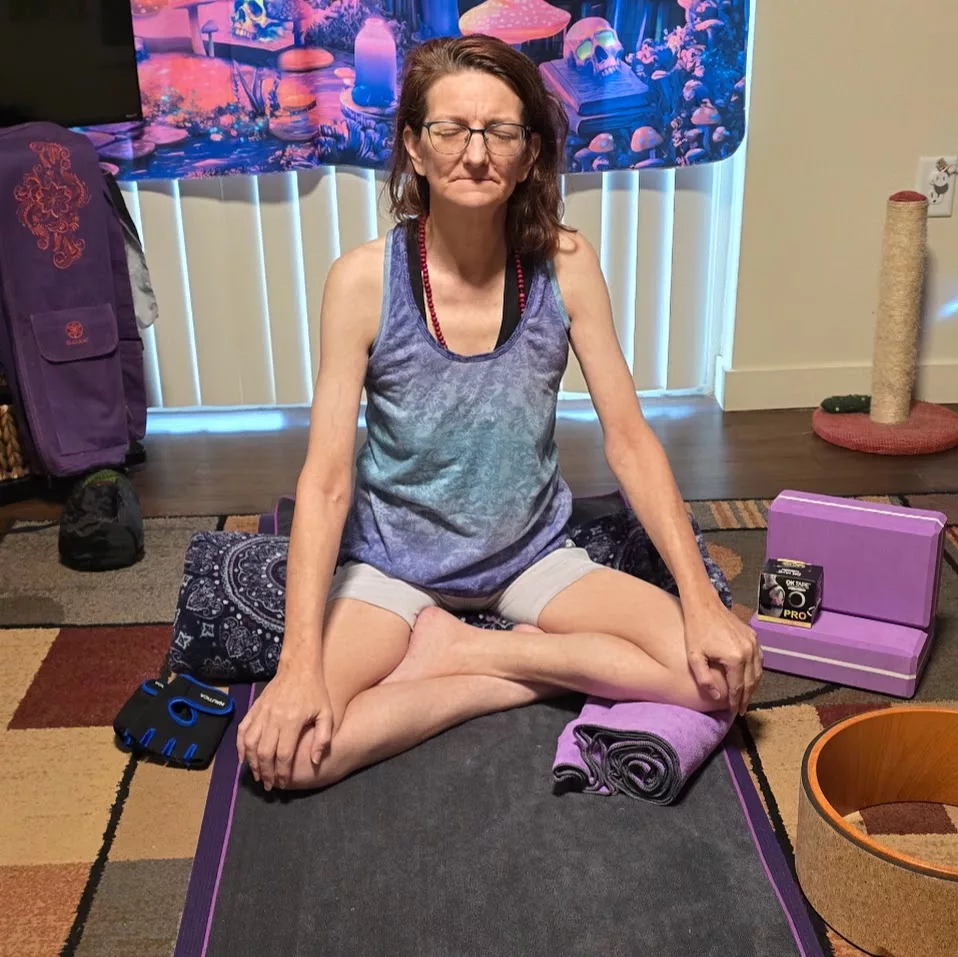 Person seated cross-legged on a yoga mat in a cozy room, surrounded by props—capturing gentle movement for chronic illness through meditation and mindful setup.