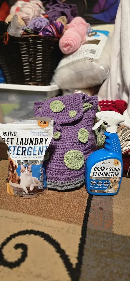 ACTIVE Pet Laundry Detergent and Odor & Stain Eliminator Spray placed on a carpeted floor beside a handmade purple crochet bag with green patches. In the background, baskets of yarn and crafting supplies suggest a cozy, pet-friendly maker space.