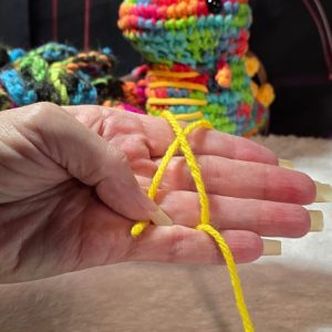 Hand demonstrating the starting loop of a magic circle with yellow yarn, shown against a soft background. Long, manicured nails and colorful plushies in the backdrop add a playful, comfort-core aesthetic—ideal for beginners learning how to make a magic circle in crochet.