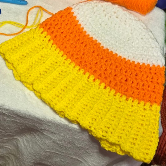 A close-up of a crochet beanie in candy corn colors with visible ribbed yellow brim, displayed on a white surface with nearby crochet tools and yarn.