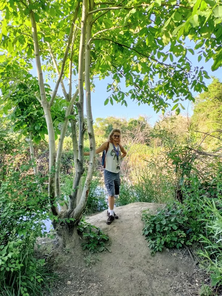 Person standing on a shaded dirt path near a tree and river, wearing hiking gear and a camera—capturing a moment of gentle movement for chronic illness in nature.