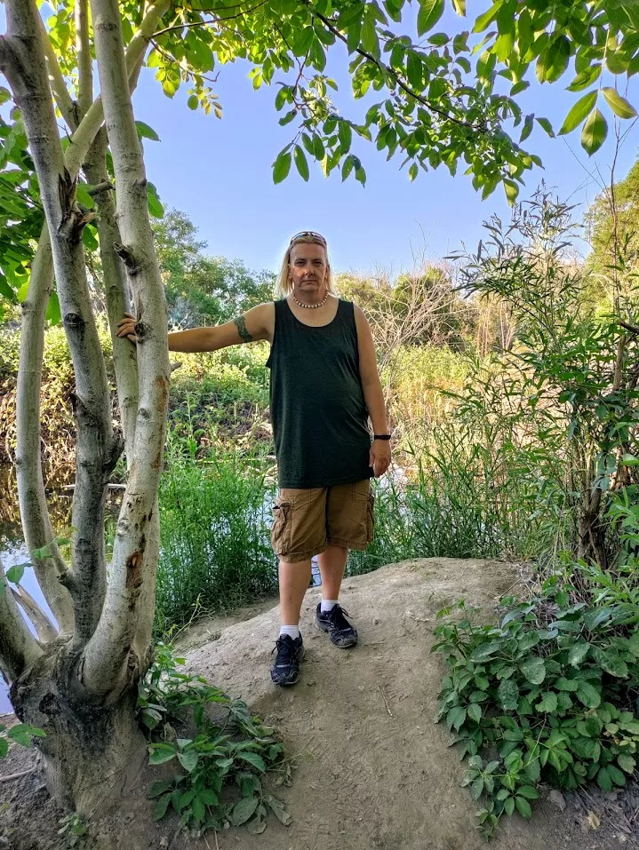 Person standing beside a tree on a sunny trail, surrounded by greenery—capturing a moment of gentle movement for chronic illness during outdoor exploration.