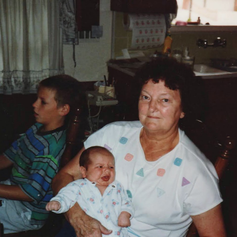 Family photo of Nana holding a baby in a patterned white outfit while a young boy plays computer games in the background. Captured in a cozy kitchen setting, the image reflects caregiving, generational connection, and the quiet moments that shaped a childhood marked by undiagnosed chronic illness.