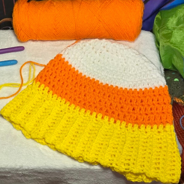 In-progress Candy Corn Beanie crochet project with white, orange, and yellow stripes, displayed beside orange yarn and crochet hooks.