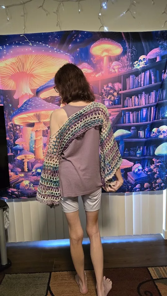 Person wearing a multicolored crocheted Bell Sleeve Fairy Shrug over a light purple tank top and white shorts, standing indoors in front of a glowing mushroom forest backdrop and bookshelf—showcasing the shrug’s whimsical drape and adjustable sleeve length in a magical, comfort-core setting.