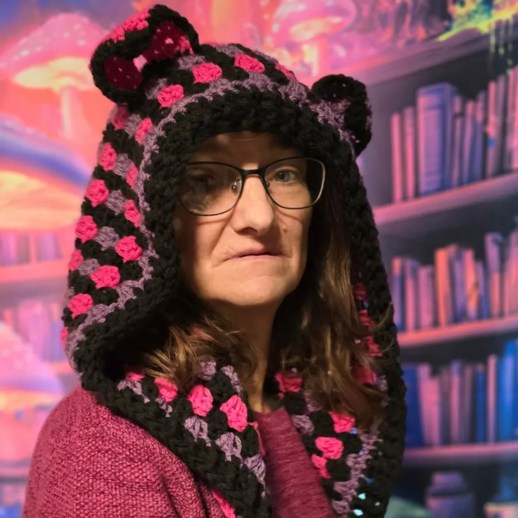 Person wearing a crocheted hooded scarf in black, pink, and purple with cat‑ear shaping, set against a vibrant fantasy‑themed background.