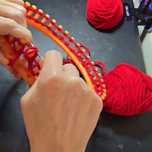 Hands wrapping red yarn around the pegs of a round knitting loom, with two balls of yarn and an “On Air” sign in the background.