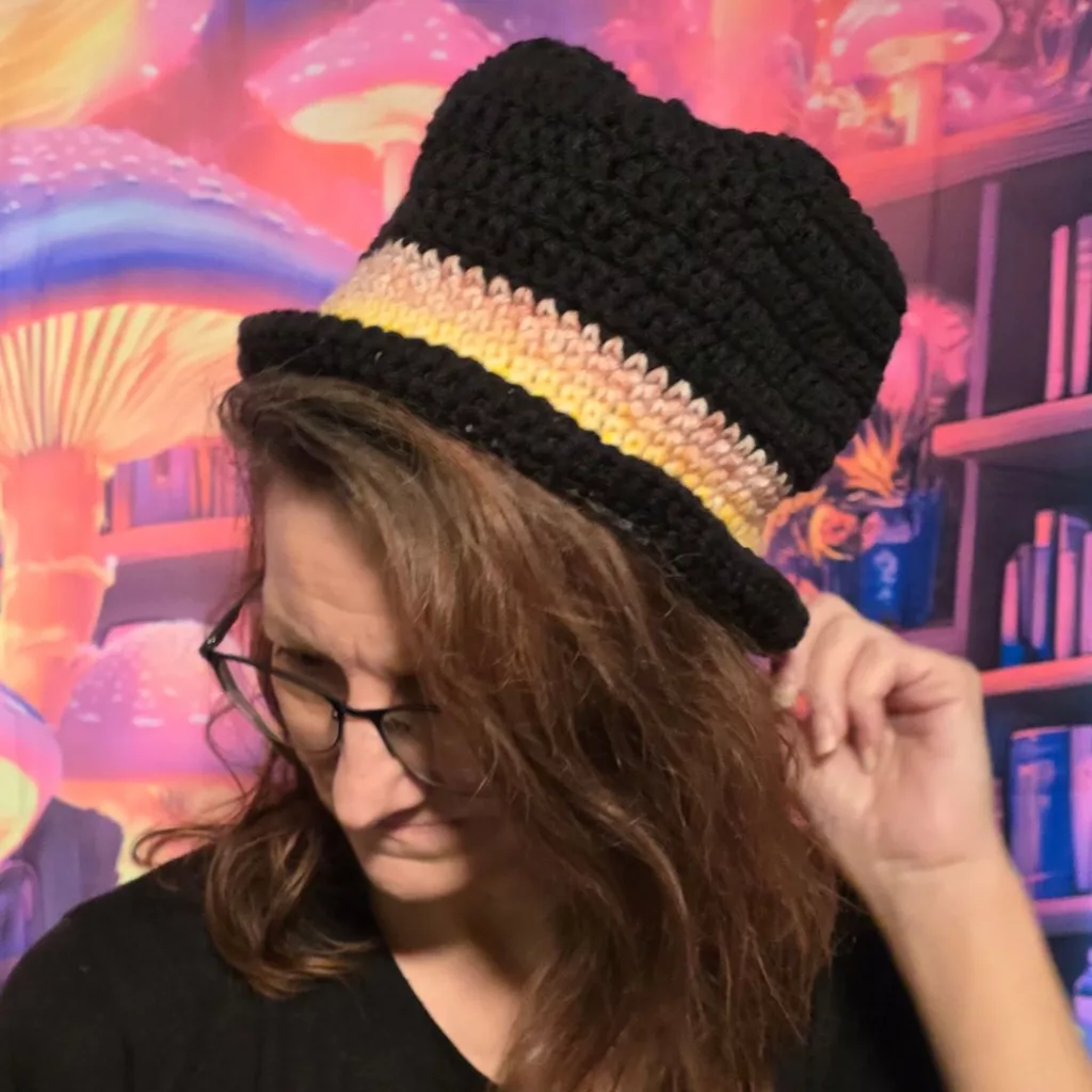 Person adjusting a black crocheted top hat with a yellow, pink, and beige band, standing before a colorful fantasy scene with glowing mushrooms and books.