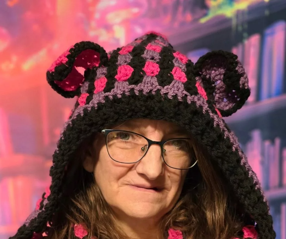 A person wearing glasses and a crochet hood with cat ears in black, purple, and pink yarn. The background is bright and abstract with colorful shapes and bookshelves.