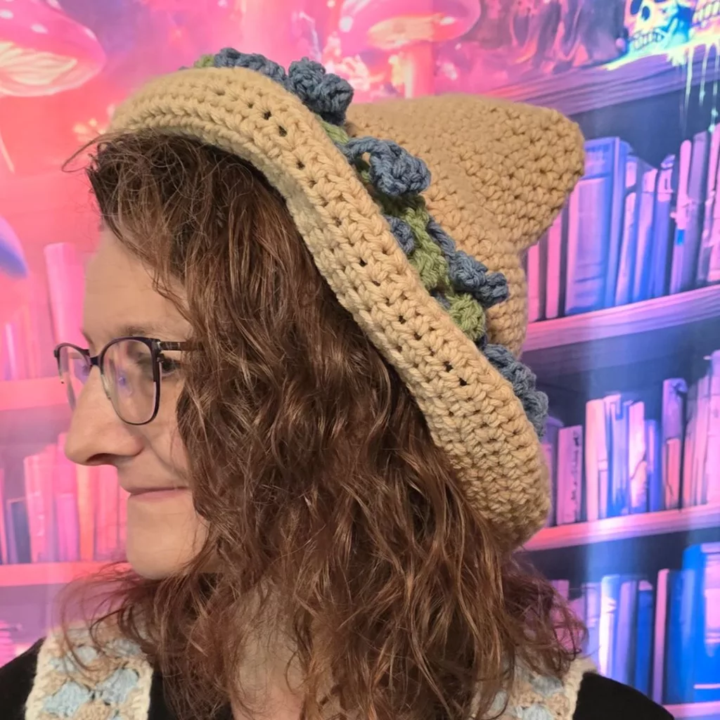 Person in profile wearing a beige crocheted hat decorated with blue and green flowers, set against a whimsical bookshelf and mushroom‑themed background.