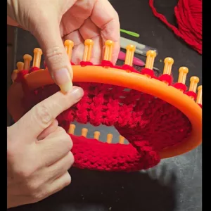 Hands wrapping bright red yarn around the pegs of a round knitting loom, creating the circular base of the hat.