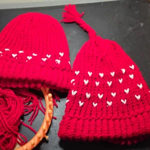 Two red loom‑knit hats with white heart motifs; one finished with a braided tassel and one still on the loom with working yarn attached.