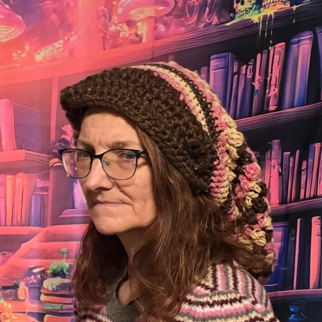 Person wearing a large multicolored crocheted slouchy hat in browns, pinks, and cream, standing in front of a magical, mushroom‑lit fantasy backdrop.
