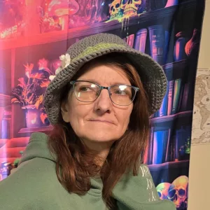 Person wearing a gray crocheted hat with a green accent and white flower, posed in front of a neon‑bright fantasy bookshelf tapestry.