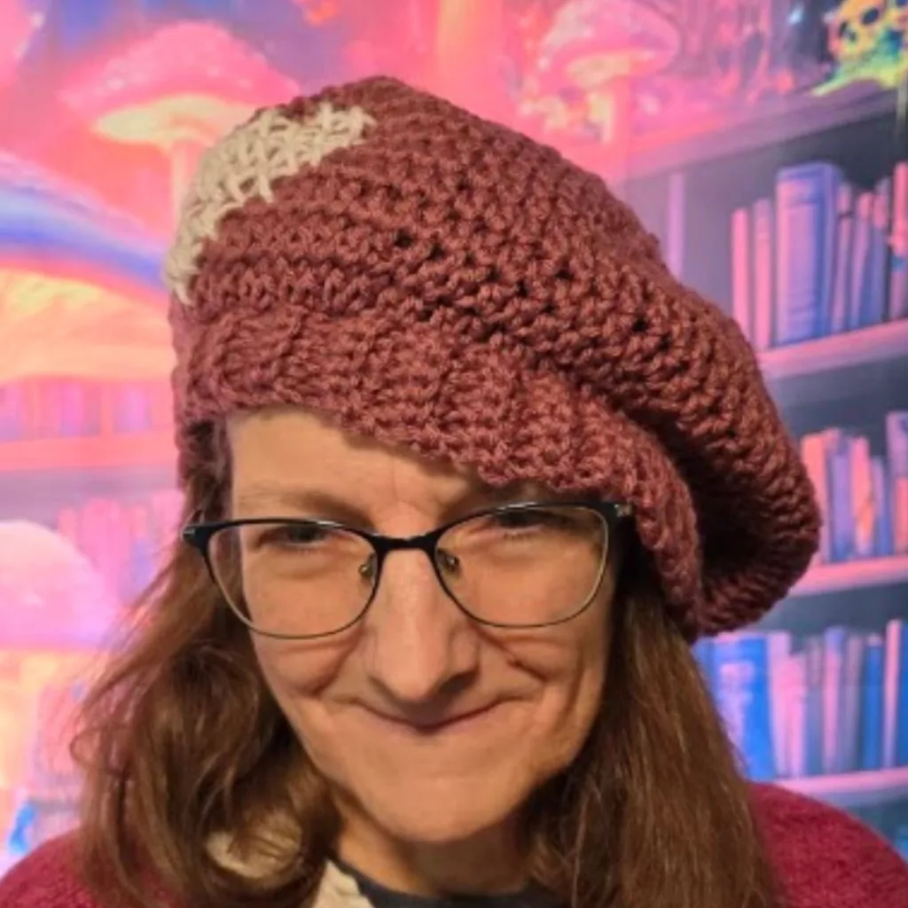 Person wearing a maroon crocheted beret with a white detail on top, smiling in front of a colorful fantasy background with glowing mushrooms and bookshelves.