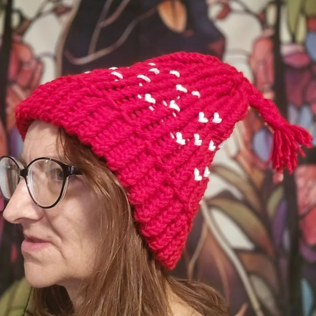 Person wearing a bright red loom‑knit hat with white heart details and a tassel, standing in front of a colorful artistic backdrop.
