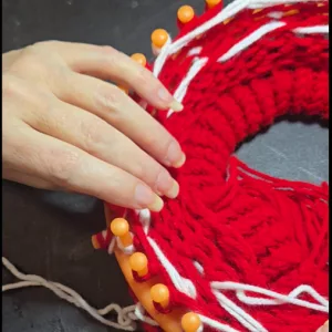 Close‑up of a round knitting loom with red and white yarn, showing a hand adjusting the stitches while working the Heartbeat Stripe pattern.