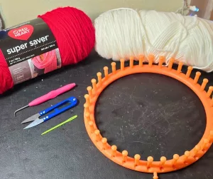 Flat lay of loom knitting supplies including red and white yarn, an orange round loom, a loom hook, scissors, and a plastic yarn needle.