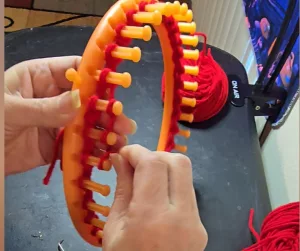 Close‑up of a person knitting on a round orange loom with red yarn, with loom tools and a ball of yarn visible in the background.