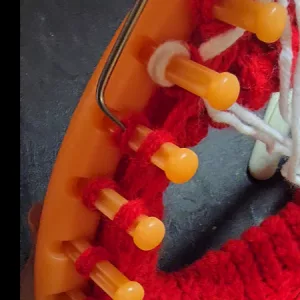 Close‑up of a knitting loom with red yarn wrapped around the pegs and a metal loom hook lifting stitches during the knitting process.