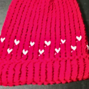 Close‑up of a red knit hat featuring a row of small white hearts worked into the fabric above the ribbed brim.
