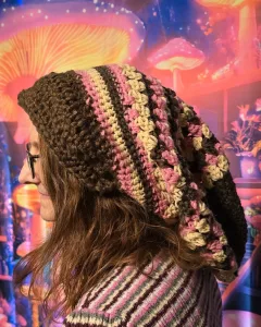 A person wearing an easy slouchy beanie crochet pattern in Neapolitan colors—brown, pink, and Buff—shown in profile against a whimsical background of glowing mushrooms in pink, orange, and purple tones.