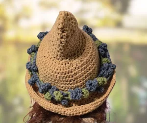A top-down view of a tan crocheted pointed hat resting on a surface. The hat has a wide, wavy brim and a decorative band made of small, blue and teal crocheted dahlias encircling the crown.
