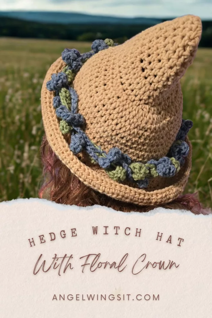 A close-up, high-angle shot of a hand-crocheted tan witch hat with a pointed top. The hat features a thick, textured floral crown made of blue and green crochet leaves and flowers wrapped around the base. The person wearing the hat has dark hair and is standing outdoors in a field.