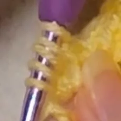 Close-up of hands crocheting a berry stitch, showing a purple crochet hook pulling a loop of orange yarn back through the same stitch.