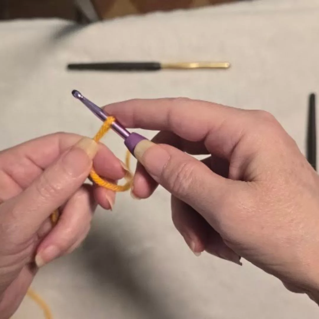 A close-up, top-down view of a person's hands performing the first step of a crochet chain stitch. The left hand holds a loop of bright yellow yarn, while the right hand holds an ergonomic crochet hook with a blue handle, positioned to yarn over and pull the strand through the loop.