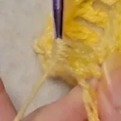 Close-up of hands crocheting a berry stitch, showing a purple crochet hook pulling the orange yarn through all loops on the hook to complete the stitch.