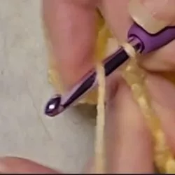 Close-up of hands crocheting a berry stitch, showing a purple crochet hook wrapping yarn over to prepare for the next loop.