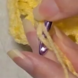 Close-up of hands crocheting a berry stitch, showing a purple crochet hook inserting into the next stitch of the orange yarn row.
