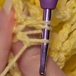 Close-up of hands crocheting a berry stitch, showing a purple crochet hook pulling a loop of orange yarn through the first loop on the hook.