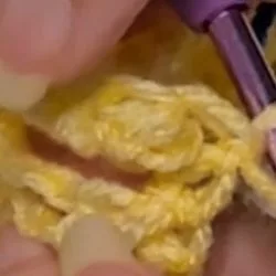 Close-up of hands crocheting a berry stitch, showing a purple crochet hook pulling a loop of orange yarn through a stitch on the row.Close-up of hands crocheting a berry stitch, showing a purple crochet hook pulling a loop of orange yarn through a stitch on the row.