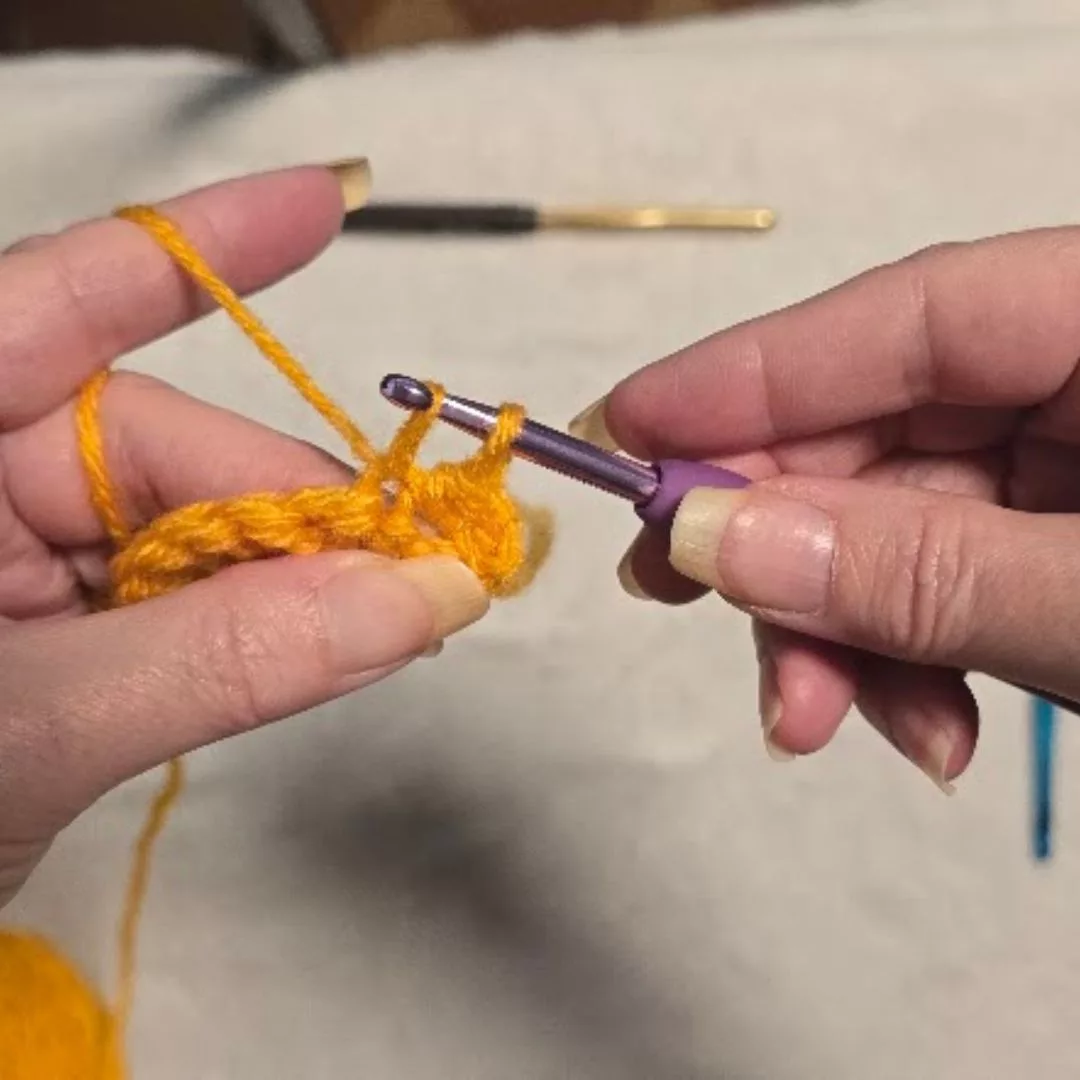 Close-up of the "pull up a loop" step for a single crochet: After the blue-handled hook is inserted into the stitch, it catches the yellow yarn and pulls it back through the stitch to the front. This leaves two loops of yarn on the hook's shaft.