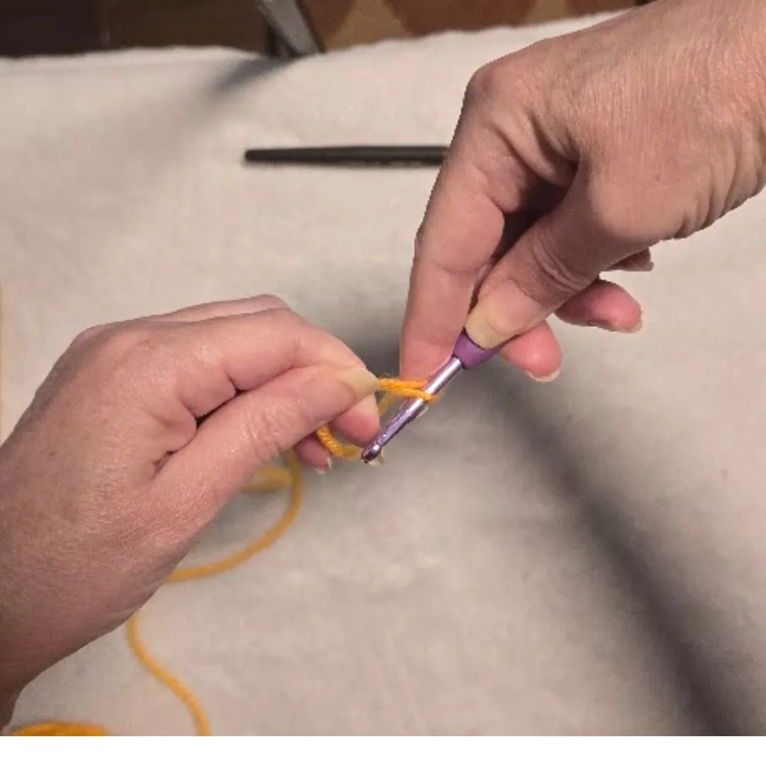 Close-up of the "pull through" step for a slip knot: The crochet hook pulls the caught strand of yellow yarn entirely through the initial loop. This creates a new adjustable loop on the hook, with the yarn ends crossing at the base to form the knot.