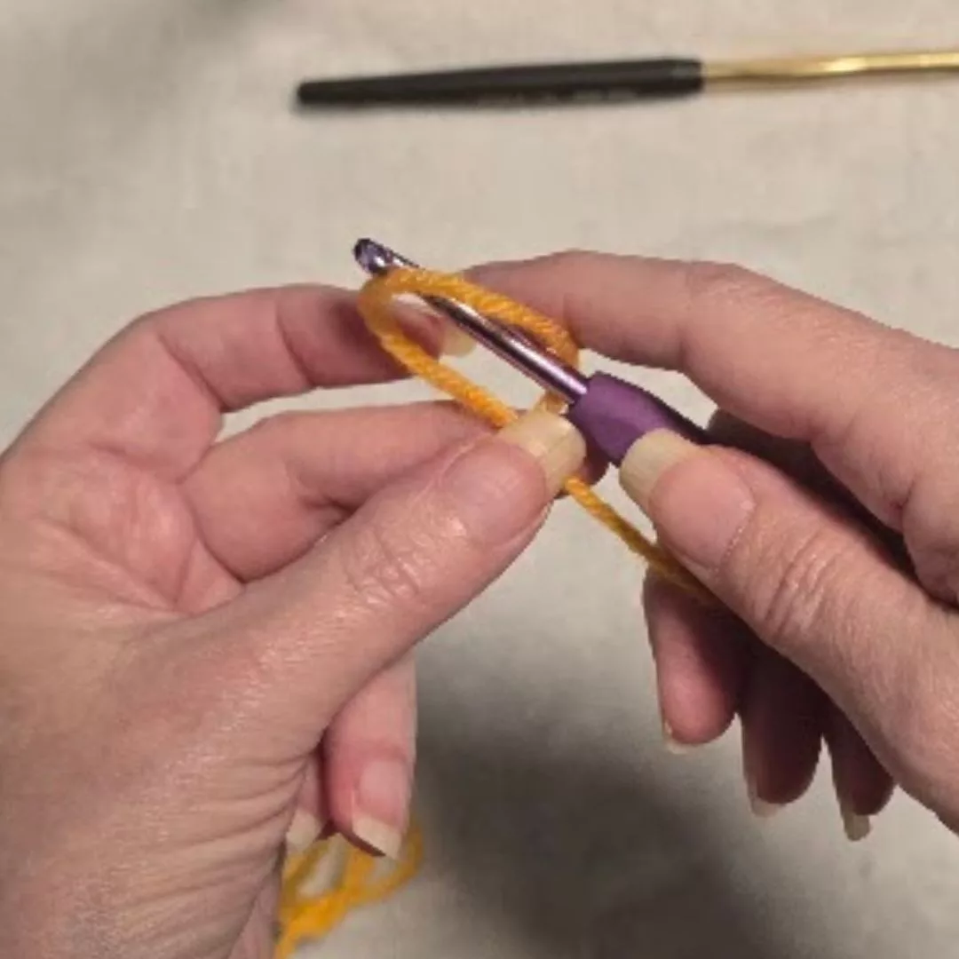 Close-up of the "insert hook" step: The blue-handled crochet hook is pushed through the center of the yarn loop. The left hand holds the loop open and steady to ensure the hook passes through without snagging the fiber.