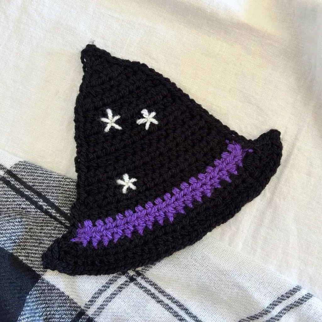 Crochet witch hat coaster with a crooked pointed crown, contrasting band and buckle.